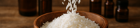 A WOODEN BOWL WITH BEUATIFUL SOY WAX FLAKES BEING POURED INTO IT WHILE THE BACKGROUND SHOWS LOTS OF OILS ON A BENCH
